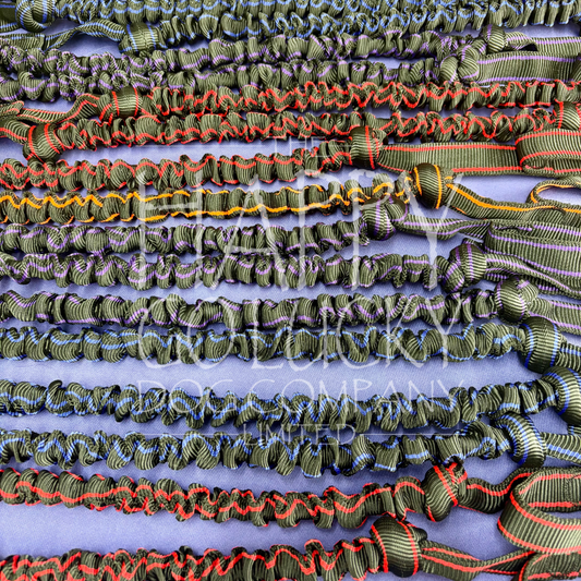 A close-up of multiple strands of black striped elastic cord trim with red, blue, and yellow accents, laid out horizontally on a light blue background. A semi-transparent watermark reading 'the happy go lucky dog company' is visible across the centre. Showing Arctic Wolf Adventure Lines